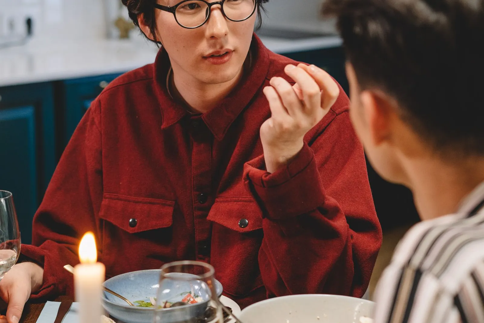 Zwei Männer im Gespräch an einem gedeckten Abendtisch mit Kerze und Weingläsern, einer mit Brille im roten Hemd, der andere im gestreiften Hemd mit dem Rücken zur Kamera