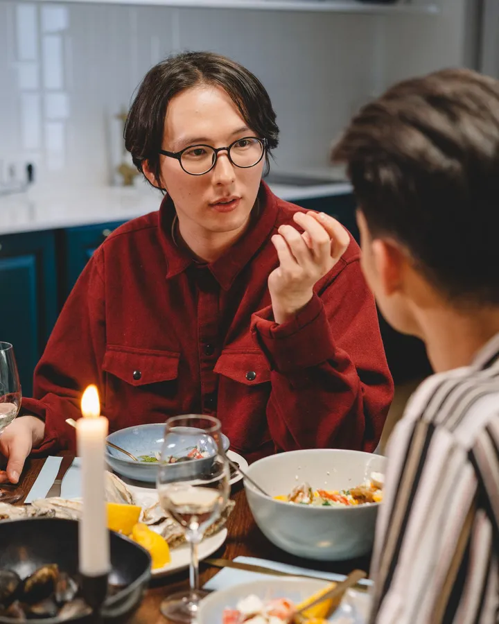 Zwei Männer im Gespräch an einem gedeckten Abendtisch mit Kerze und Weingläsern, einer mit Brille im roten Hemd, der andere im gestreiften Hemd mit dem Rücken zur Kamera