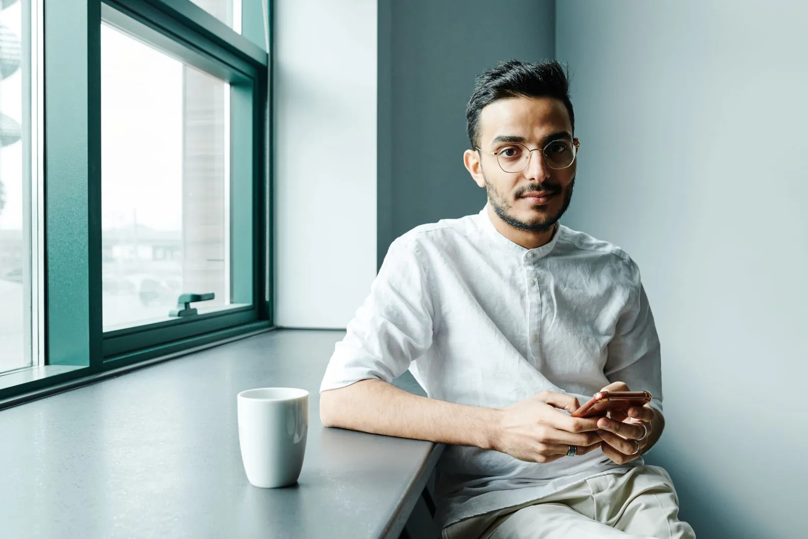 Mann mit Brille und weißem Leinenhemd sitzt allein am Fenstertresen eines hellen Cafés, Handy in der Hand, Kaffeebecher neben sich