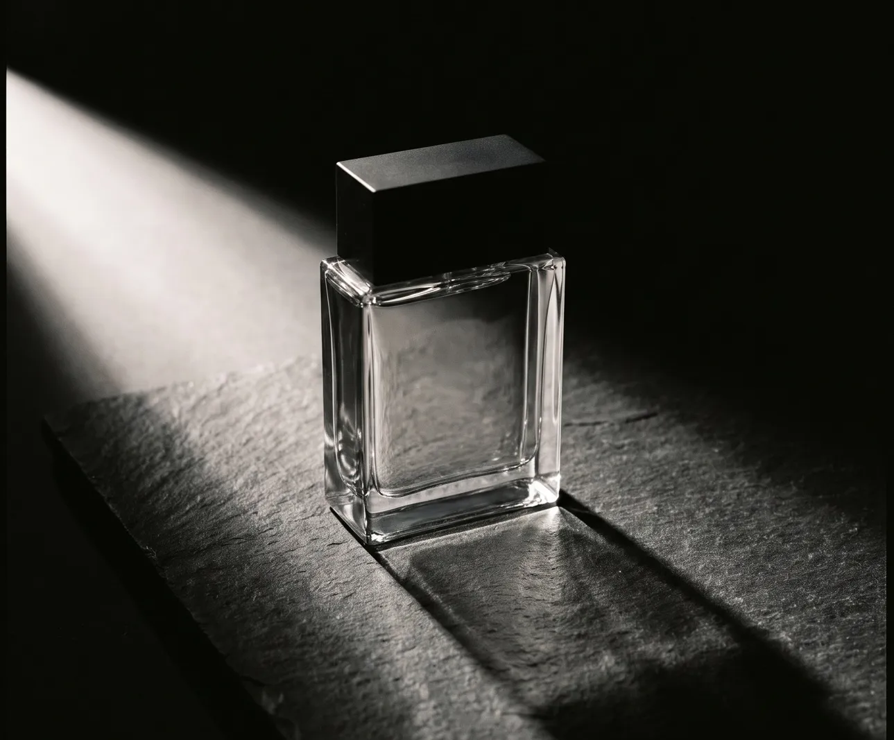 Unbeschriftete eckige Glasflasche mit schwarzer Kappe auf einer Schieferplatte, ein Lichtstreifen wirft einen langen, harten Schatten