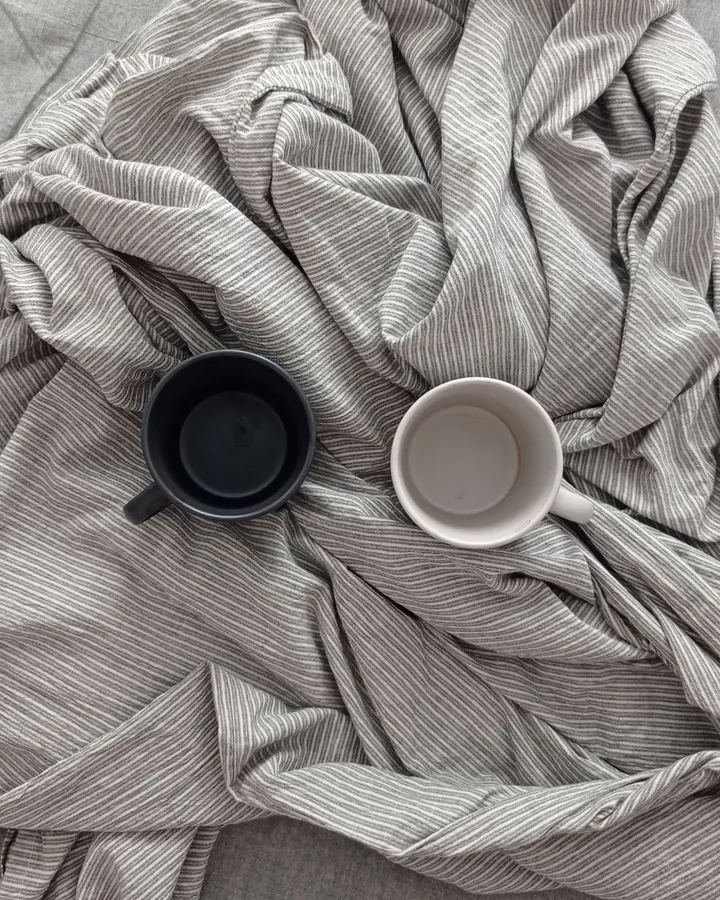 Zwei leere Kaffeebecher, einer schwarz, einer weiß, auf zerwühlter grau gestreifter Bettwäsche, von oben fotografiert
