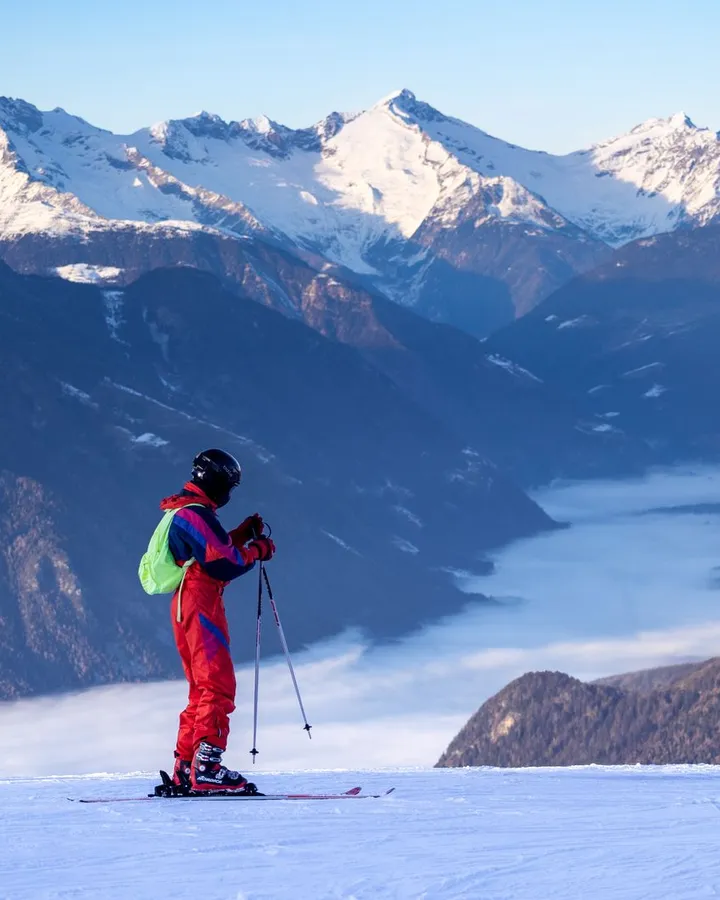 Skifahrer im roten Retro-Skianzug steht auf der Piste und blickt über ein nebelgefülltes Alpental auf verschneite Gipfel