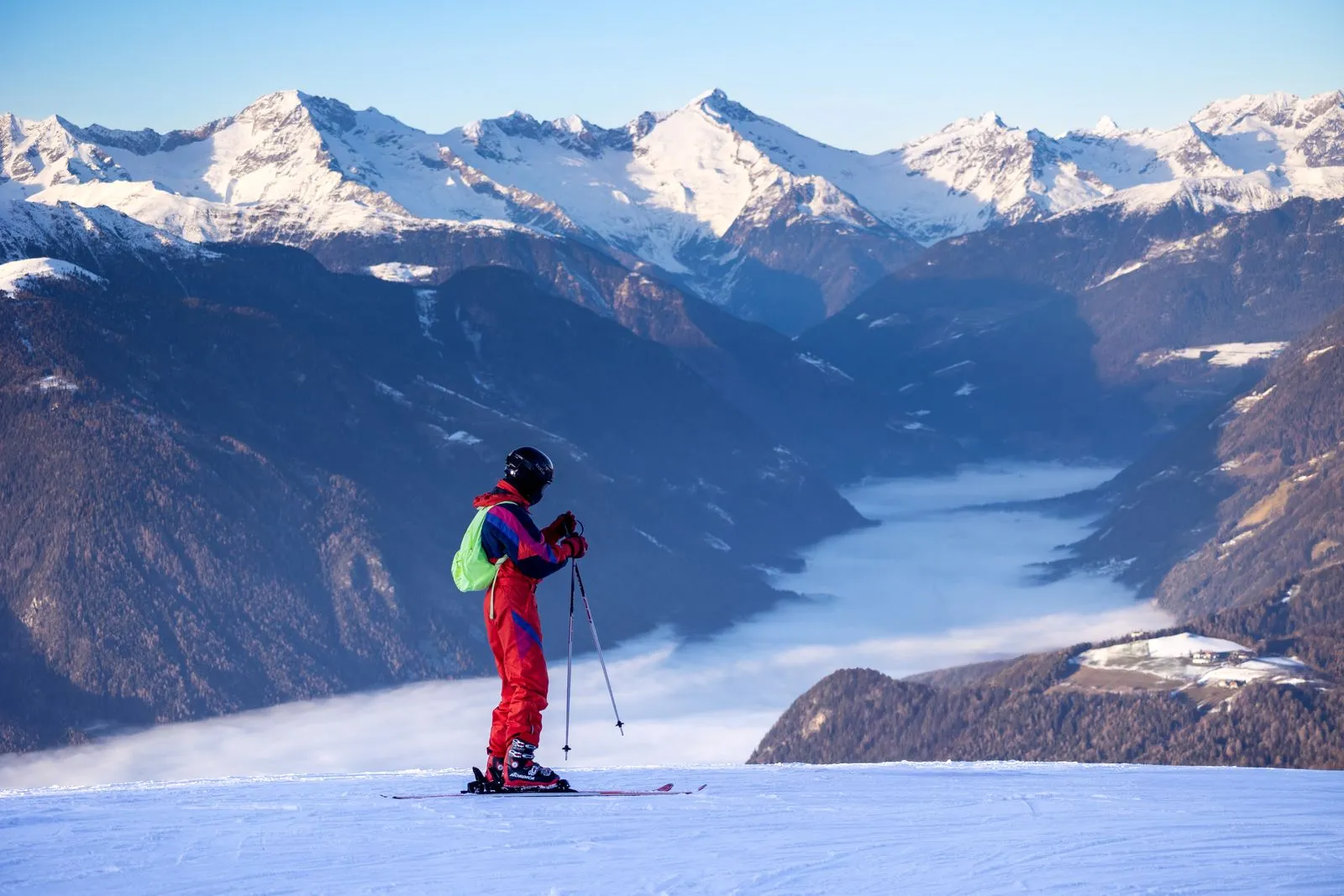Skifahrer im roten Retro-Skianzug steht auf der Piste und blickt über ein nebelgefülltes Alpental auf verschneite Gipfel