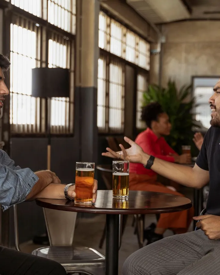 Zwei Männer sitzen sich in einer hellen Bar mit hohen Sprossenfenstern an einem kleinen runden Tisch gegenüber, vor ihnen zwei Gläser Bier, einer erzählt mit ausgestreckter Hand, der andere hört lächelnd zu