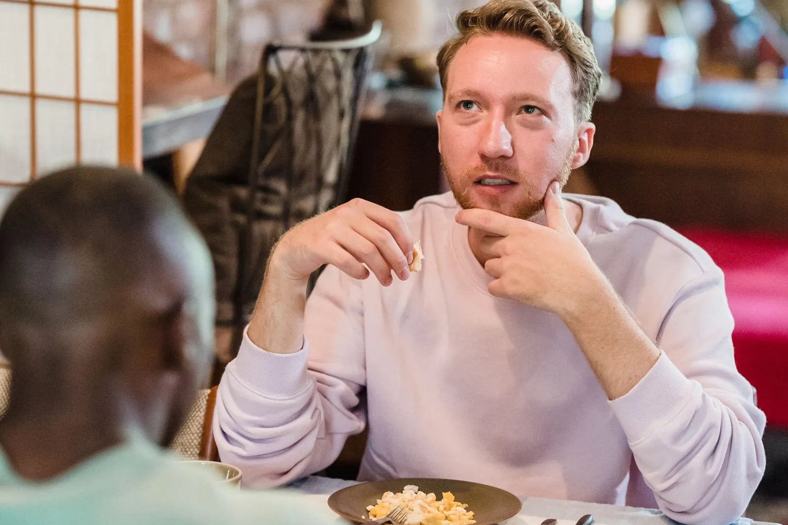 Zwei Männer beim Frühstück am Holztisch, einer im fliederfarbenen Pullover blickt nachdenklich zur Seite, der andere sitzt mit dem Rücken zur Kamera, davor Teller und Kaffeetasse