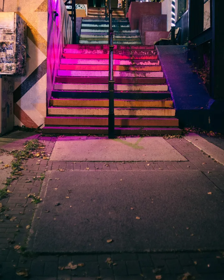 Leere Treppe mit abgewetzten Regenbogenstufen bei Nacht, magentafarbenes Licht, Herbstlaub auf dem Gehweg
